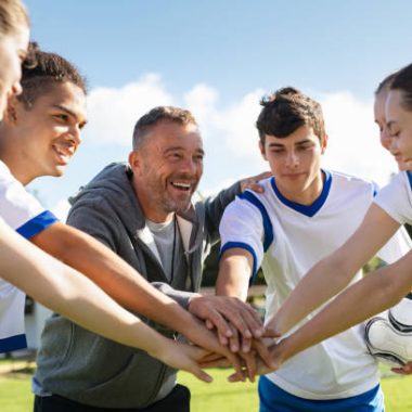 Happy coach and high school soccer team with their hands stacked in football court. Young football players stacking hands together. Mature man coach encourages his students to do their best during the sport match.