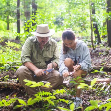 A father teaching his young daughter wilderness survival, bushcraft and knife skills in a forest.